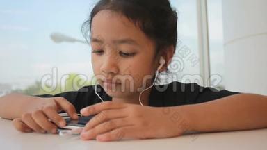 亚洲小女孩使用智能手机，耳朵耳机，坐在玻璃窗附近